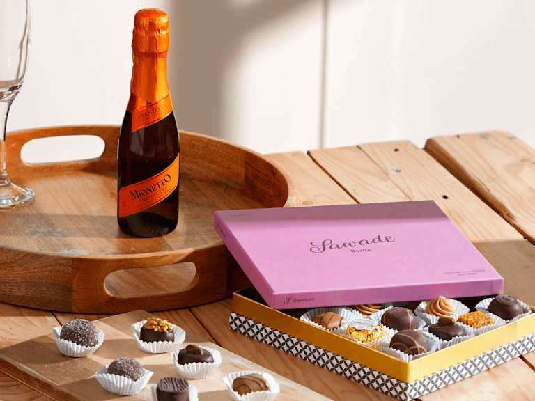 Eine rosafarbene, edle Box mit einer gemischten Auswahl an Pralinen von Sawade Berlin liegt halb geöffnet auf dem Tisch, dahinter ist eine Piccolo-Flasche Prosecco von Mionetto zu sehen.