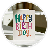 Greeting card with colourful hand-lettered "Happy Birthday" text in rainbow colours on a cream background, laid on a wooden desk.
