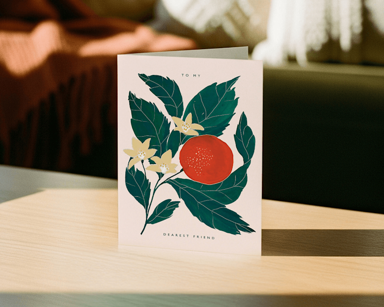 A greetings card illustrated with an orange and some foliage sits on a work surface.