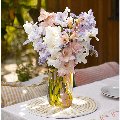 White and pink gladioli bouquet with purple statice in a glass vase, set on a table with striped cushions in dappled sunlight.