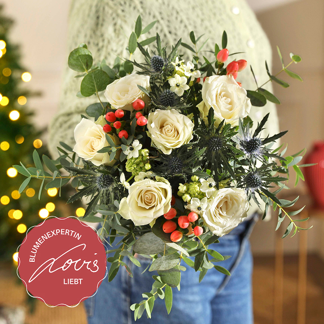 Zarter Winterstrauß mit cremeweißen Rosen, stahlblauem Distelkopf, roten Beeren und Eukalyptusgrün, gehalten vor einem beleuchteten Tannenbaum.