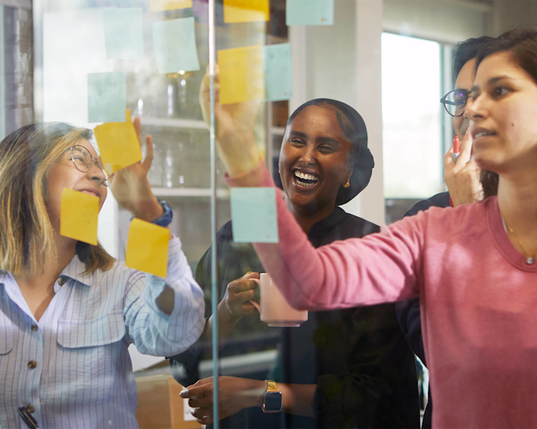 Three women are laughing and smiling as they stick post-it notes on a wall. Two are medium-skinned, and one is dark-skinned and wearing a headscarf.