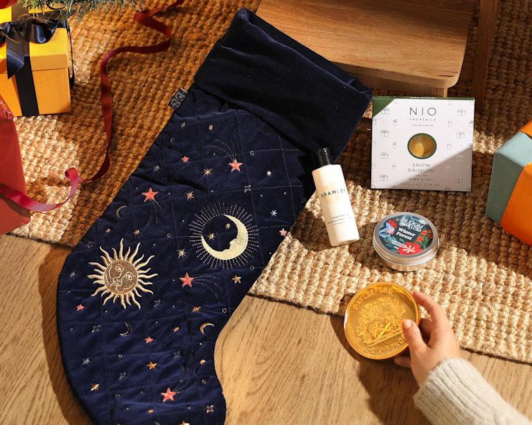 An Elizabeth Scarlett stocking lying on a rug, embroidered with moons and stars. With a letterbox cocktail, a chocolate coin, a candle, and a hand cream.
