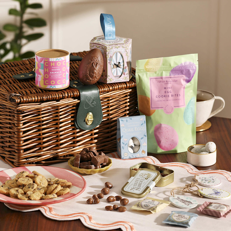 A traditional wicker picnic hamper overflowing with Easter treats. Features an embossed chocolate egg, "Mini Egg Cookie Bites", and praline 'quail' eggs. A mix of smooth chocolate, crunchy biscuits, and ornate packaging creates an indulgent feel.