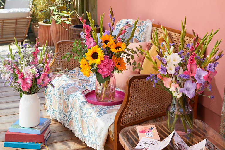 A sunny outdoor scene featuring three vibrant bouquets. One bouquet, with sunflowers and pink hydrangeas, sits on a wooden bench with patterned cushions. Two other bouquets, bursting with colourful gladioli and stocks, stand nearby.