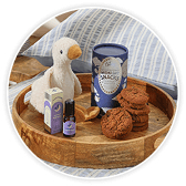 Wooden tray holding a plush toy duck, a striped biscuit tin, a jar of biscuits, and two small bottles, styled as a gift hamper.