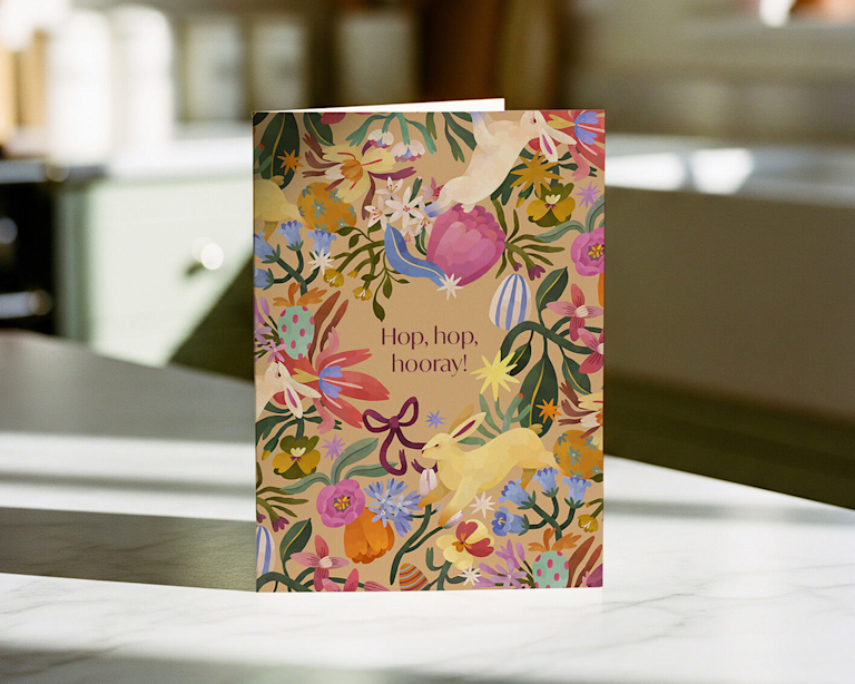 A vibrant Easter greeting card featuring the message "Hop, hop, hooray!" surrounded by a whimsical illustration of leaping bunnies and colourful spring flowers. The matte-finish card uses a joyful palette of pink, yellow, and blue.