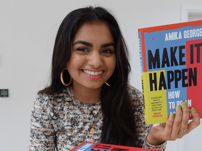 Amika, a light-brown-skinned woman with long black hair and big gold hoops, smiles and holds up her book Make It Happen