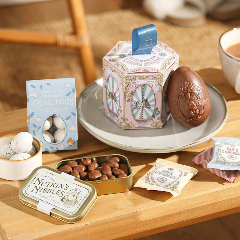 A whimsical Easter chocolate selection featuring a carousel-themed gift box, an embossed milk chocolate egg, and "Nutkin’s Nibbles" tin and a variety of smooth chocolate, crunchy praline eggs.