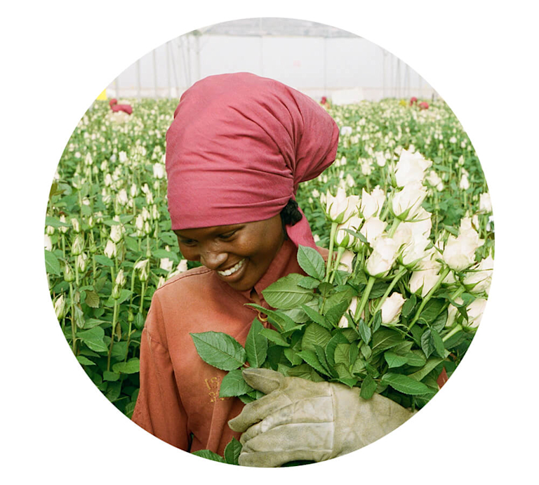 A smiling dark-skinned woman in a headscarf, overalls and gardening gloves cradles an armful of white roses.