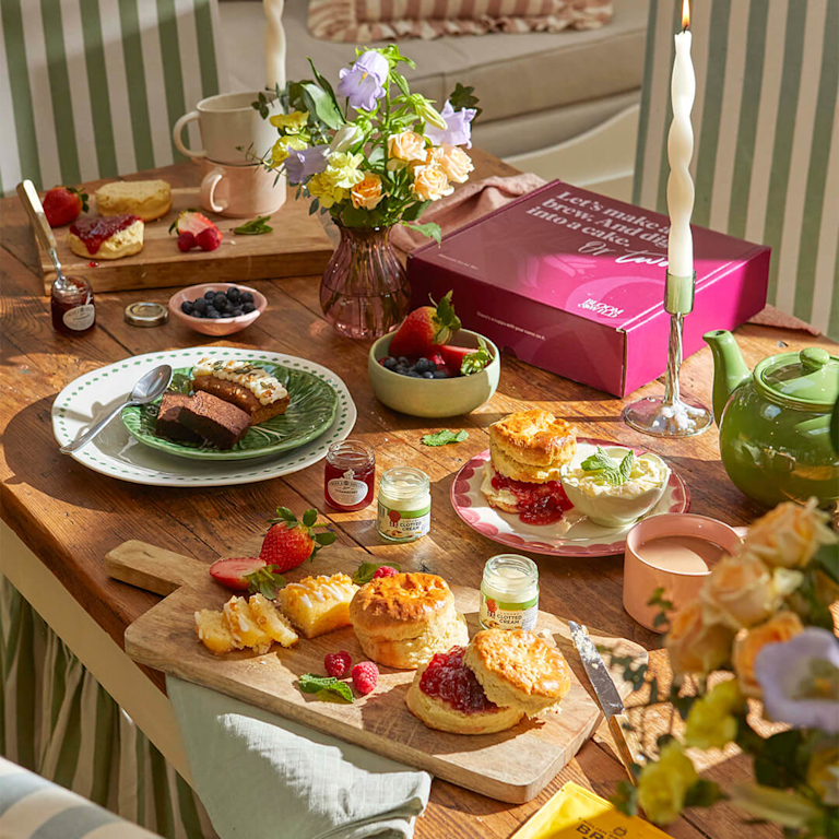 A luxurious afternoon tea spread featuring golden, crumbly scones with strawberry jam and clotted cream, alongside chocolate brownies and lemon cake. A pastel bouquet in a glass vase and steaming tea create a fragrant, celebratory table scene.