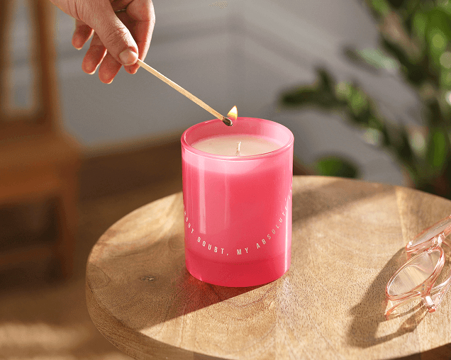 A hand lighting a pink glass jar candle with a long wooden match on a round wooden side table.