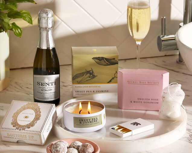 A luxury gift set on a marble tray featuring a small bottle of champagne, a glass of bubbly, a lit "Sweet Pea & Jasmine" candle, and chocolate truffles.