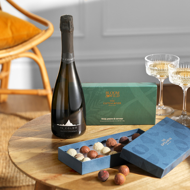 A celebration scene is set on a light wooden round table, featuring a bottle of Il Colmo Prosecco, an open blue box of assorted chocolates with some scattered on the table, and two stemmed glasses. The overall mood suggests a special occasion.