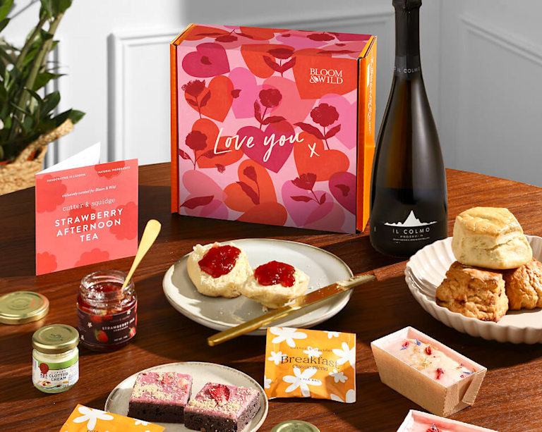 A Valentine's Day afternoon tea gift set on a wooden table featuring a "Love You" heart-patterned box, a bottle of Prosecco, scones with jam and clotted cream, chocolate brownies, mini loaf cakes, and tea sachets.