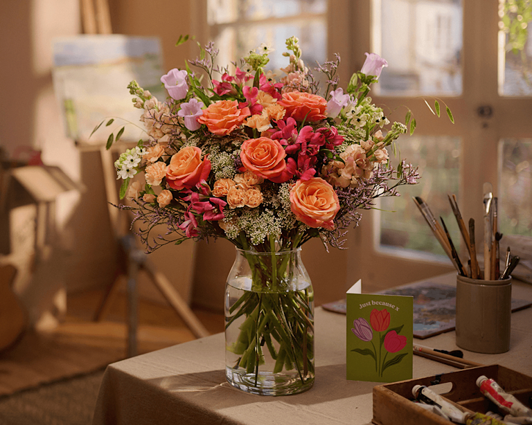 The 'Helen' bouquet by Bloom & Wild featuring a mix of orange and pink roses, lilac campanula and ammi, sits in a glass vase upon a surface in an art studio.