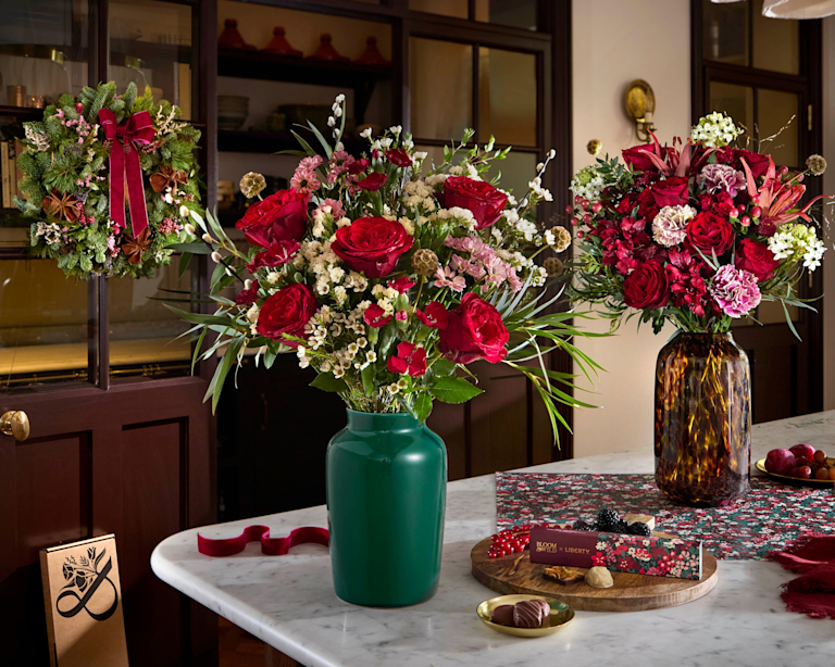 Red and pink Liberty Christmas bouquets in a green vase and a tortoiseshell vase on a marble counter, with a box of Liberty-print truffles and a festive wreath on the cabinet behind.