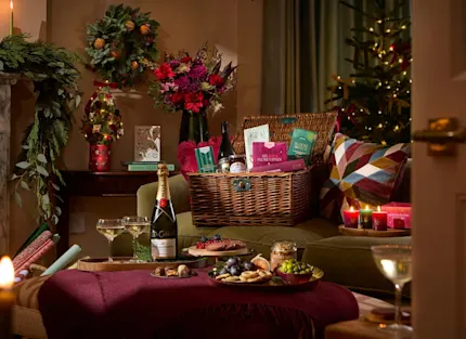 A cosy living room at Christmas with a wicker hamper of festive treats on a green sofa, a bottle of Champagne and glasses, lit red candles, a grazing board on a burgundy footstool and a decorated tree.
