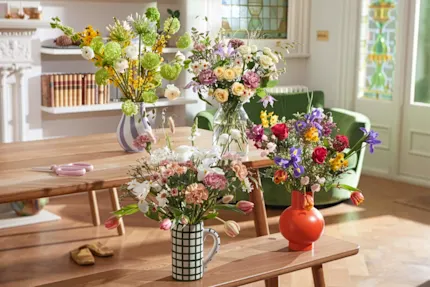 Four blossoming bouquets, with a variety of springtime stems, sit in vases across a dining room table and bench. A pair of pink floristry scissors rest on the table.