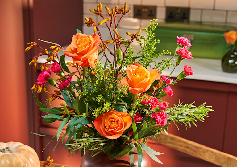 Bright autumnal bouquet of orange roses, pink carnations and green foliage in a brown vase on wooden kitchen table.