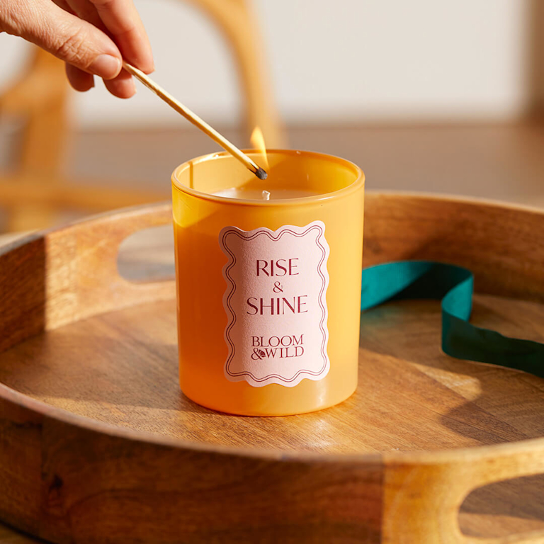 A person lighting a "Rise & Shine" scented candle in a warm amber glass jar, sat on a wooden tray.