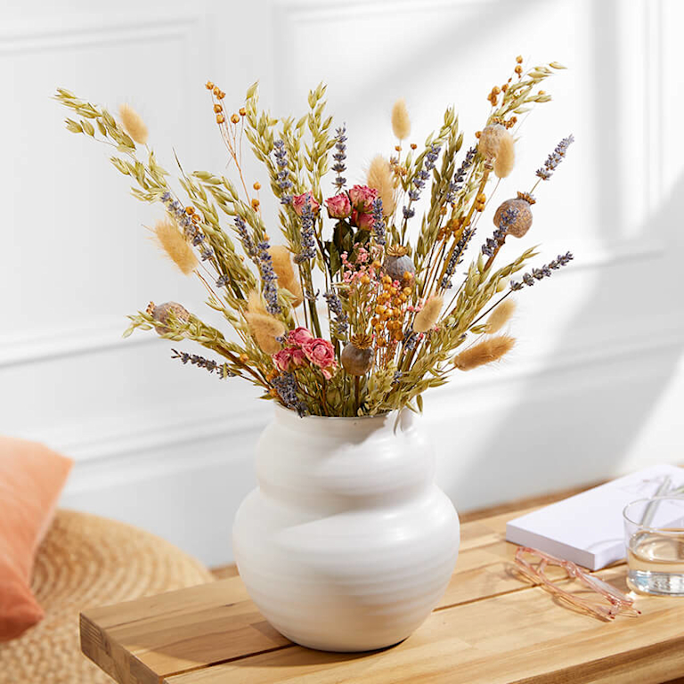 A vase of earthy-toned dried stems are placed in a white vase. Homely objects are placed around it.
