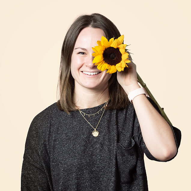 Emily is a white woman in her thirties with long brown hair. She smiles and holds up a sunflower to her face.