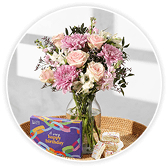 Pink and white roses with lilac and blush flowers and greenery in a glass vase, beside a purple Happy Birthday chocolate box on a wooden table.