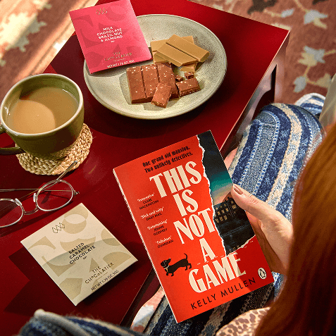 Person holding Kelly Mullen's novel This Is Not a Game, by a red side table with a mug of tea, glasses, a salted caramel chocolate bar, a milk chocolate bar and a plate of chocolate pieces.