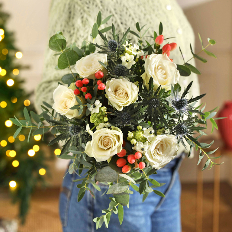 Zarter Winterstrauß mit cremeweißen Rosen, stahlblauem Distelkopf, roten Beeren und Eukalyptusgrün, gehalten vor einem beleuchteten Tannenbaum.