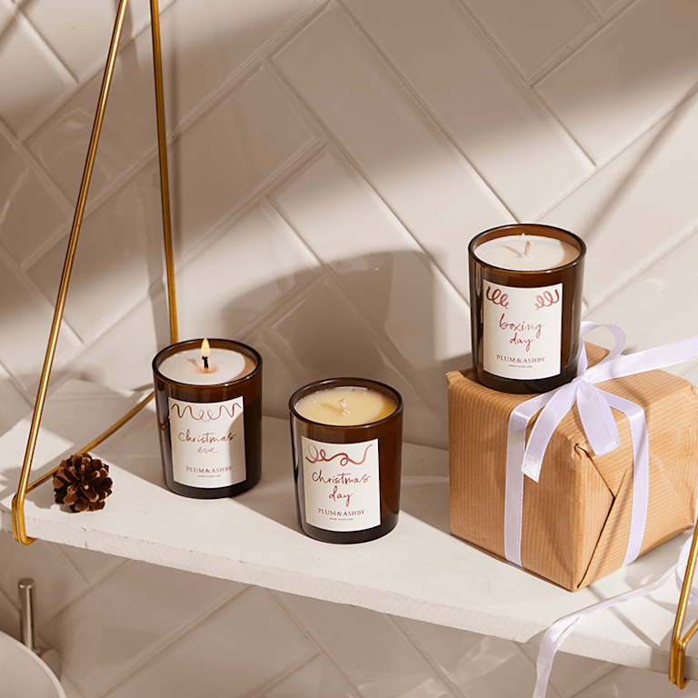 A trio of Plum & Ashby candles in brown glass jars, lined up on a shelf in a stylishly-tiled bathroom.