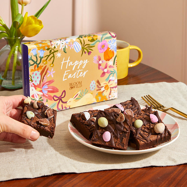 Indulgent, fudgy chocolate brownies topped with crunchy mini eggs. The "Happy Easter" gift box in the background features a vibrant floral illustration.