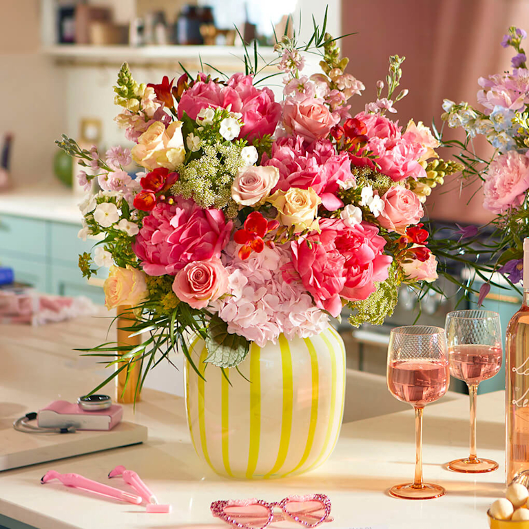 Lush bouquet of pink peonies, roses, and colorful blooms in a cream and yellow striped vase. Two pink drinks in stemmed glasses and pink sunglasses sit on a white counter. A bottle is partially visible. Kitchen setting in the background.