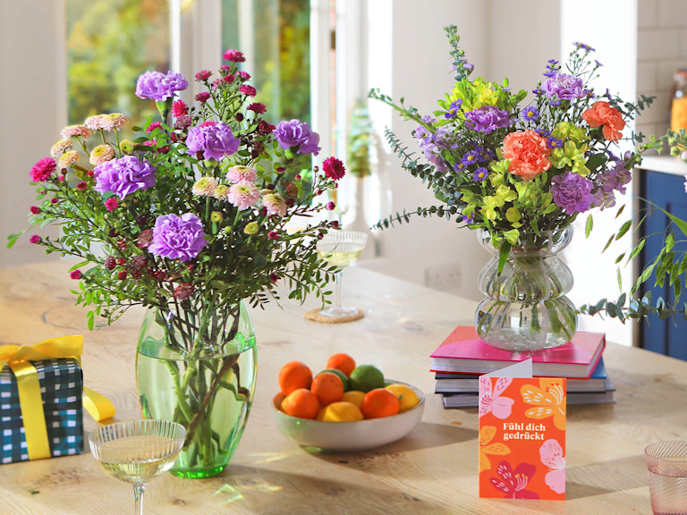 Zwei blühend schöne Blumensträuße stehen gemeinsam mit einer Grußkarte auf dem Esstisch