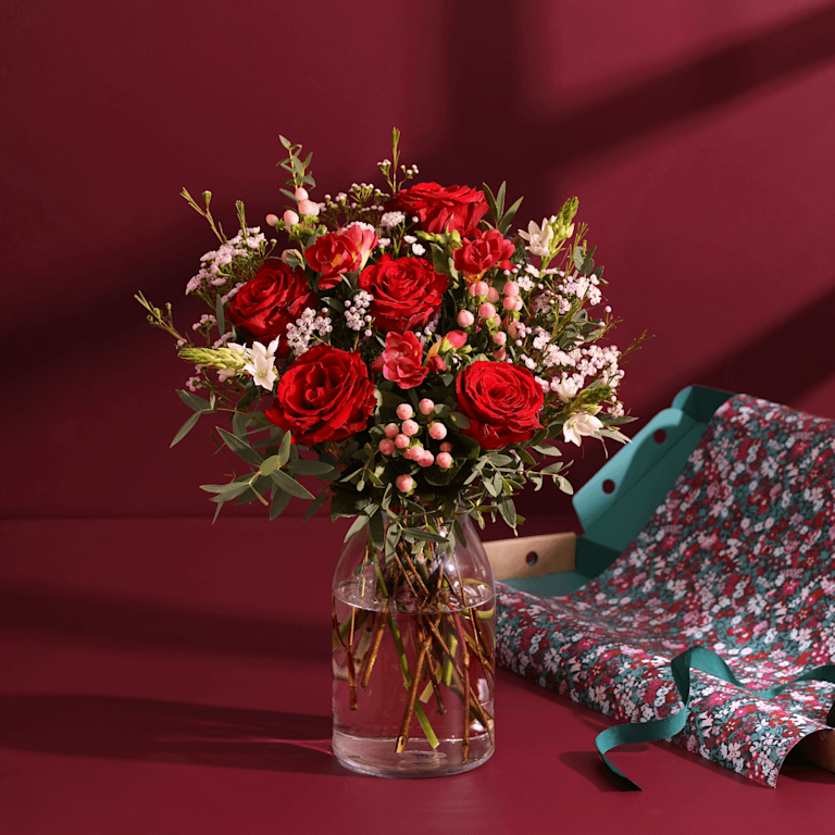 An arrangement of roses, snapdragons and white hypericum berries. An open box, lined with a Liberty print, is positioned behind.