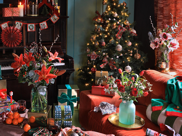 A festive decorated living room, featuring a twinkling Christmas tree, winter bouquets in vases, and wrapped gifts.