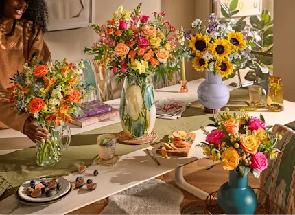 A woman arranges four colorful bouquets—orange roses, mixed pink and yellow blooms, sunflowers, and pink-orange roses—in varied vases on a dining table styled with fruit, books, and a runner in a sunlit room.