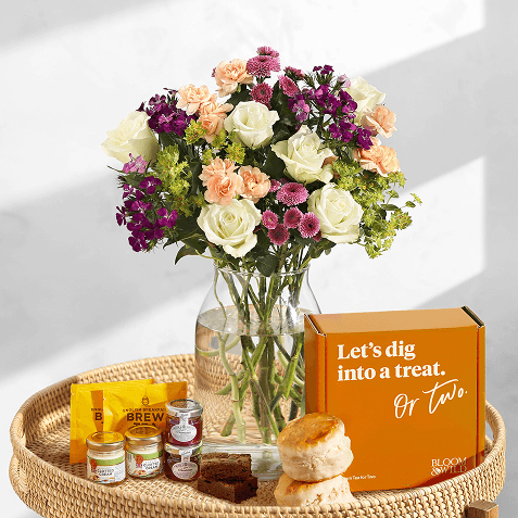 White roses, peach carnations, purple and pink blooms and green foliage in a glass vase on a rattan tray, with scones, brownies, jam jars, tea bags and an orange treat box.