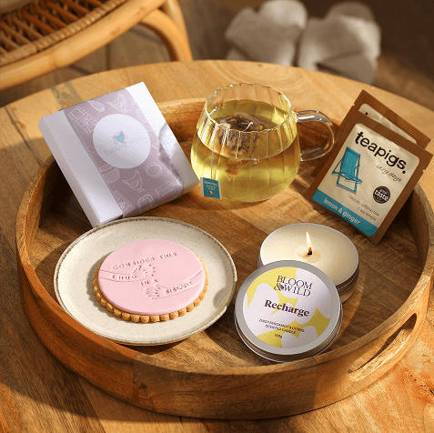 Wooden tray holding a pink iced biscuit on a plate, a glass cup of tea, lemon and ginger tea sachets, a lit Bloom & Wild Recharge candle and a small wrapped gift.