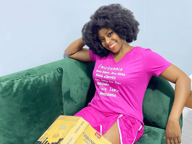 Chimamanda, a dark-skinned woman with a centre-parted natural afro, reclines on a sofa and smiles. A book rests in her lap.