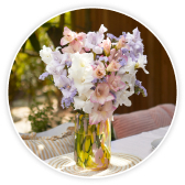 Pastel bouquet of white and lilac gladioli and roses in a glass vase on an outdoor table.