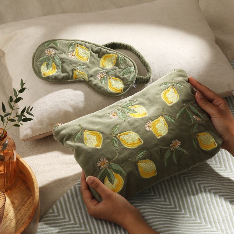 A person gently holds a soft, olive-green velvet pouch embroidered with bright yellow lemons and delicate white flowers with green leaves. Beside it, on a textured cream surface, lies a matching eye mask.