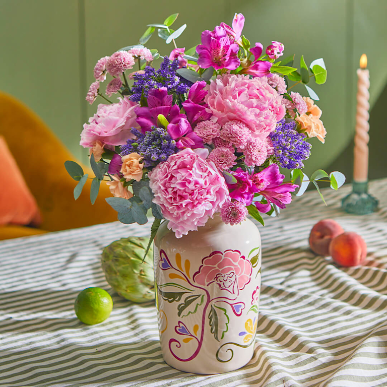 Bright yellow round vase with light pink peonies, purple flowers, and eucalyptus leaves. On a white surface with pink sunglasses. Blurred background with a person in yellow.