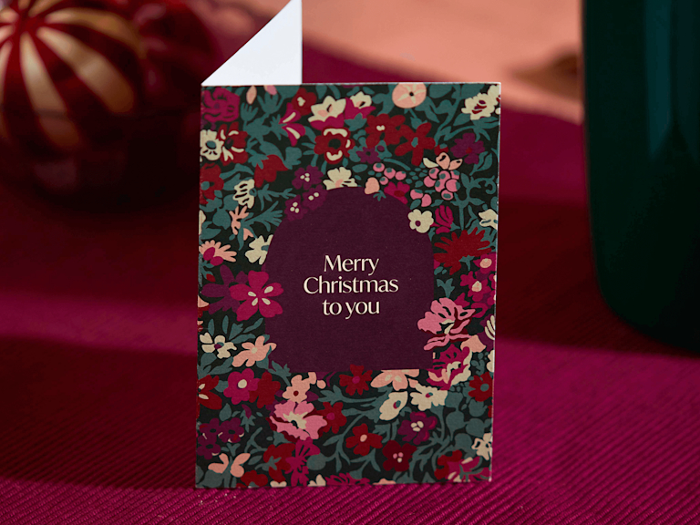 A Christmas greetings card, decorated with a Liberty print, on a rich maroon coloured background.