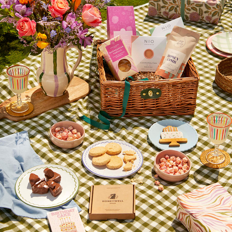 Sunny picnic scene on a gingham rug. A wicker hamper overflows with treats from sustainable brands like NIO Cocktails and Honeywell Bakes. Beside it, a jug of vibrant pink roses and purple wildflowers creates a joyful, celebratory atmosphere.