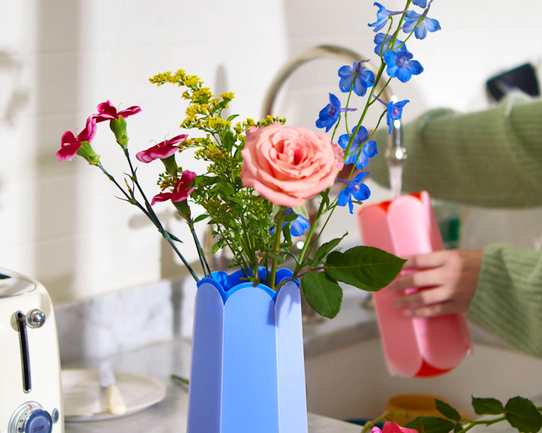 Several stems sit pretty in a letterbox vase. Behind, a woman fills up another letterbox vase with water at a sink.