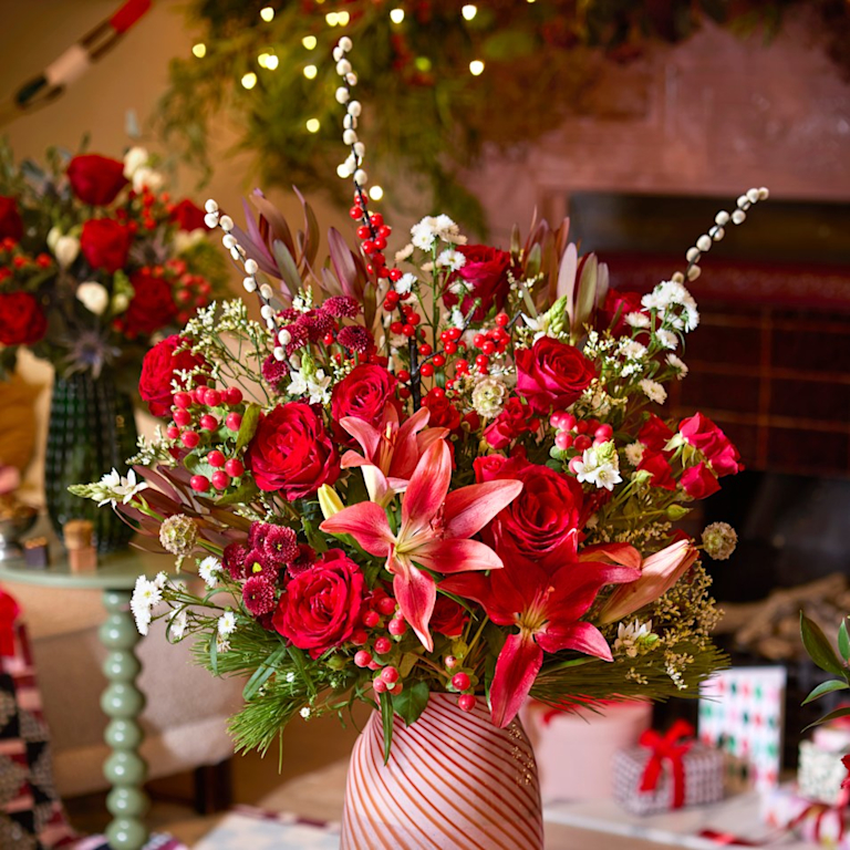 A showstopping bouquet of red roses, red lilies, white blossom and berried winter foliage in a red and white striped vase.
