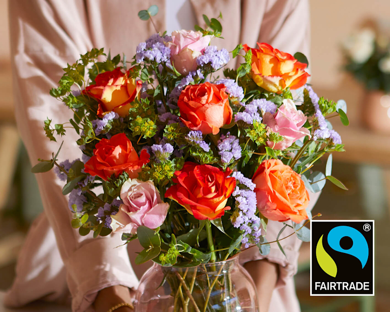 A close-up of a vibrant Fairtrade bouquet held in a glass vase. Fiery orange roses contrast with soft pink blooms and textured purple statice. The Fairtrade logo is visible, highlighting the ethical and sustainable sourcing of these joyful flowers.