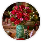 Red roses and winter foliage in a green glass vase, with twinkling Christmas lights behind.