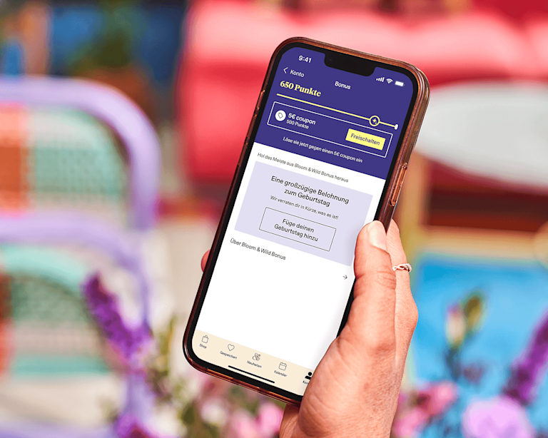 A hand holds a smartphone. The screen shows the Bloom & Wild Rewards member's area.
